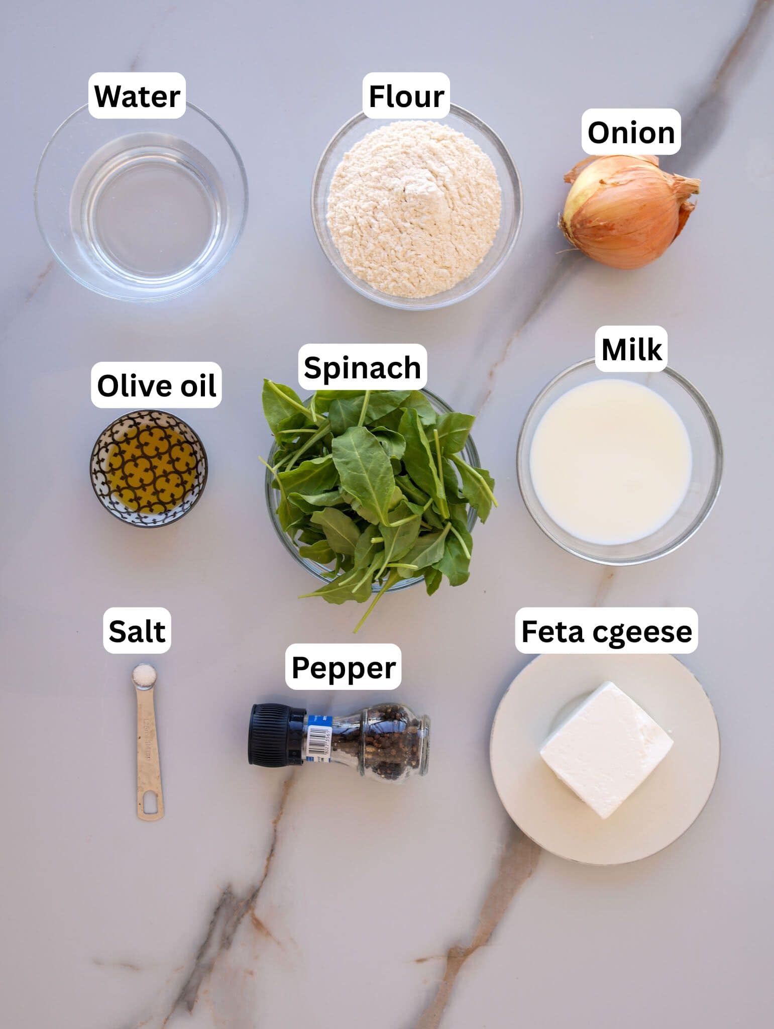 Overhead view of labeled ingredients on a counter: water, flour, onion, olive oil, spinach, milk, salt, pepper, and feta cheese. Each item is in a separate container or bowl.