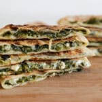 Stacked slices of flatbread filled with sautéed greens and white cheese, displayed on a wooden surface, showing the vibrant green and creamy filling inside the golden brown pastry.