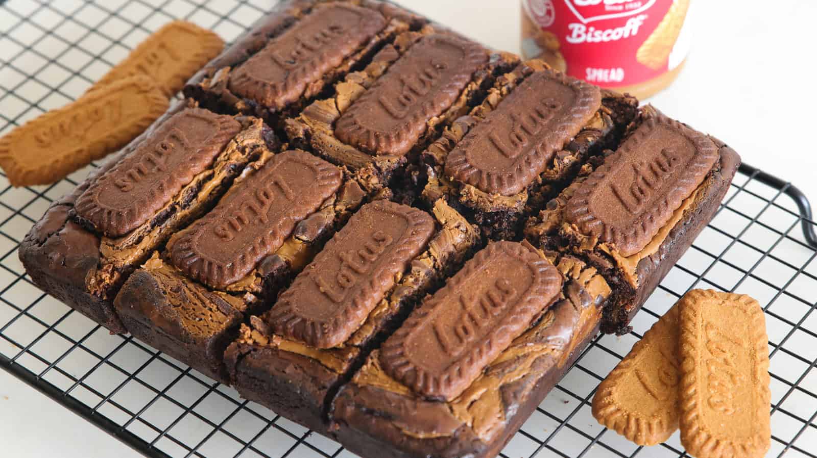 A batch of chocolate brownies topped with Lotus Biscoff cookies, cut into squares, cooling on a wire rack. A jar of Lotus Biscoff spread is visible in the background, next to some additional Biscoff cookies.