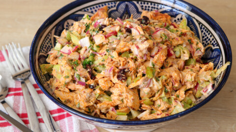 A decorative blue and white bowl filled with a colorful chicken salad featuring shredded chicken, diced vegetables, and dark dried fruits, garnished with fresh herbs. The bowl is placed on a wooden surface next to a red and white checkered cloth with forks and spoons.