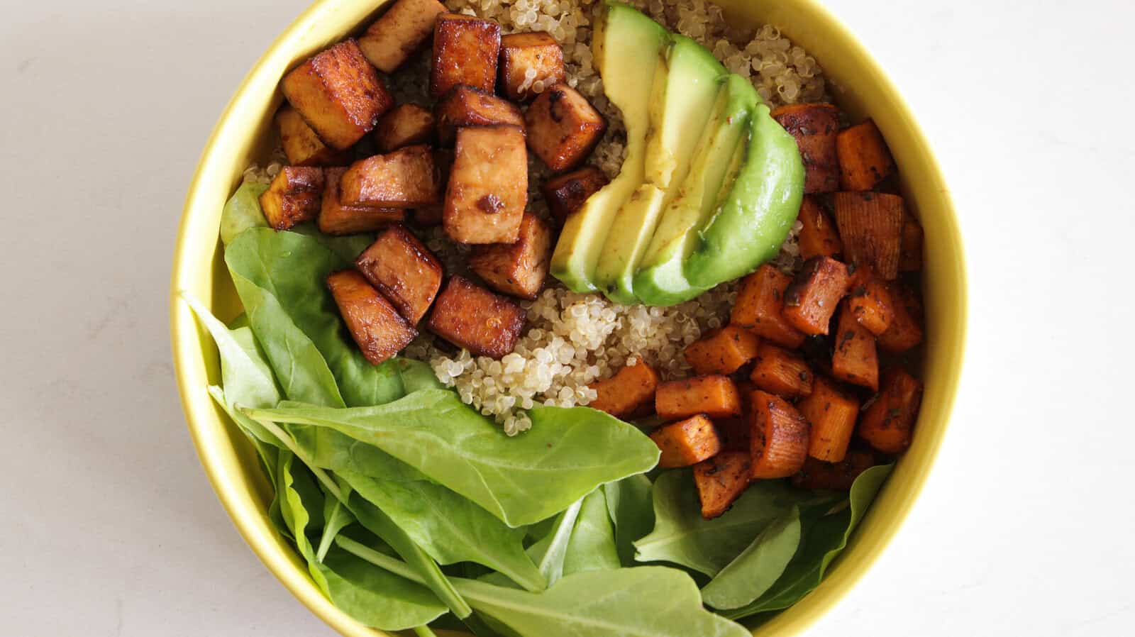 A vibrant bowl of food containing quinoa, diced tofu, sliced avocado, cubed sweet potatoes, and fresh spinach leaves, all arranged in sections. The ingredients are colorful and neatly organized, creating an appetizing and healthy meal.