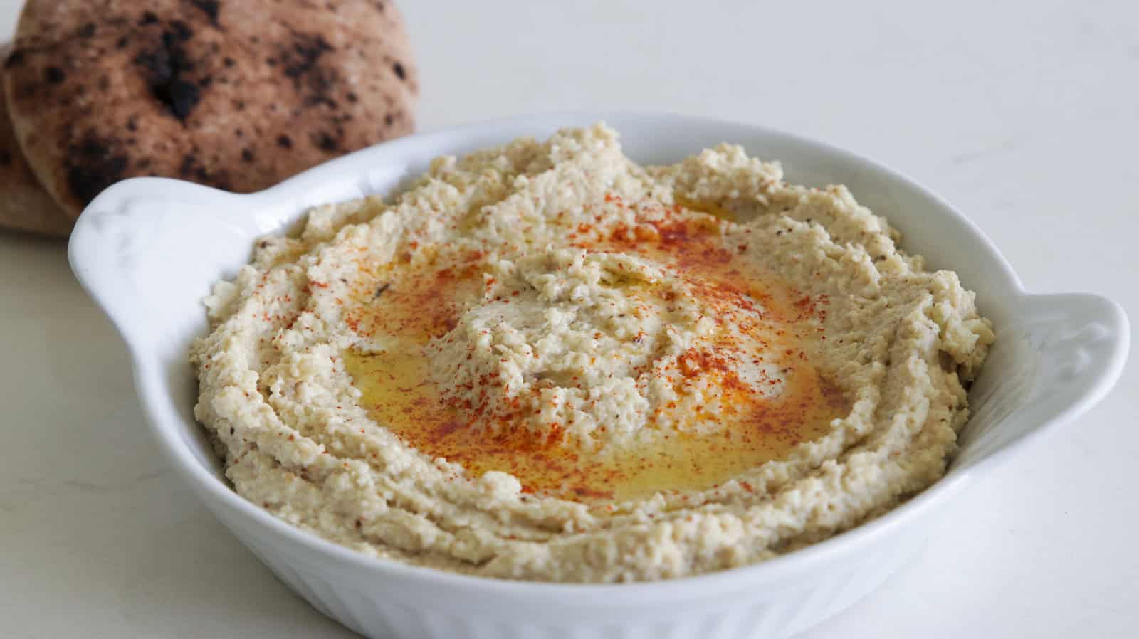 A white dish filled with hummus topped with olive oil and paprika, served with a side of pita bread in the background and a bowl of sliced carrots to the right.