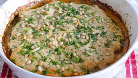 A baked Siniya dish in a white casserole topped with a golden layer, sprinkled with chopped herbs and pine nuts, resting on a red and white checkered cloth.