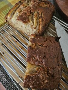 A loaf of banana bread, partially sliced, rests on a wire cooling rack. A knife lies nearby, and a piece of the bread is separated from the main loaf, showing its moist, textured interior.