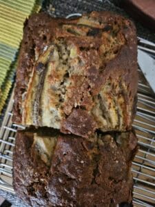 A loaf of homemade banana bread with a cracked golden-brown crust rests on a cooling rack, with a knife nearby and a slice partially cut. A piece of banana is visible baked into the top.