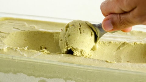 A hand is shown scooping pistachio ice cream from a rectangular container. The text "PISTACHIO ICE CREAM" is displayed at the top of the image. The ice cream has a creamy, light green color typical of pistachio flavor.