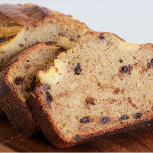 Close-up of a sliced banana bread loaf on a wooden cutting board. The bread is moist, with visible banana pieces and chocolate chips throughout. The crust is golden brown, contrasting with the lighter, speckled interior. The background is neutral and out of focus.