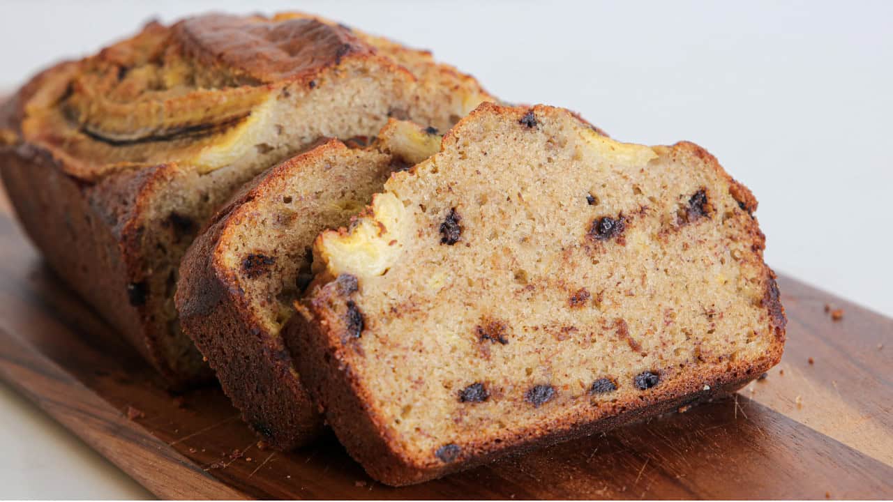 Close-up of a sliced banana bread loaf on a wooden cutting board. The bread is moist, with visible banana pieces and chocolate chips throughout. The crust is golden brown, contrasting with the lighter, speckled interior. The background is neutral and out of focus.