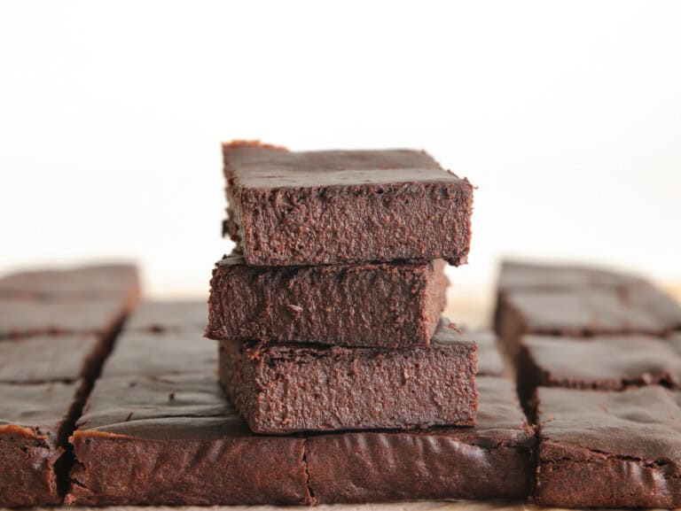 A stack of three chocolate brownies sits on top of a larger array of brownies, all cut into squares. The brownies have a rich, dense texture and a dark chocolate color. The background is out of focus, emphasizing the dessert.