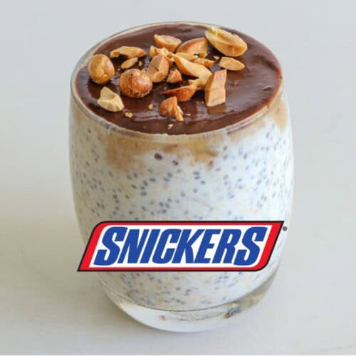 A glass filled with overnight oats topped with a layer of chocolate and sprinkled with chopped nuts. Text on the image reads "SNICKERS" in blue and red letters, and "OVERNIGHT OATS" in white letters above the glass.