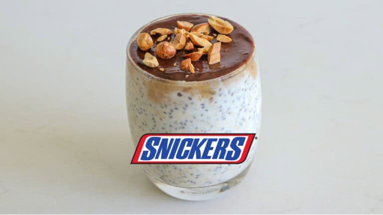 A glass filled with overnight oats topped with a layer of chocolate and sprinkled with chopped nuts. Text on the image reads "SNICKERS" in blue and red letters, and "OVERNIGHT OATS" in white letters above the glass.