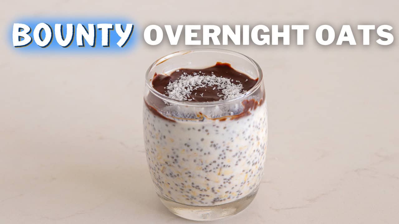 A glass of overnight oats topped with a layer of chocolate and sprinkled with coconut flakes on a white surface. Bold white text above the glass reads "Bounty Overnight Oats.