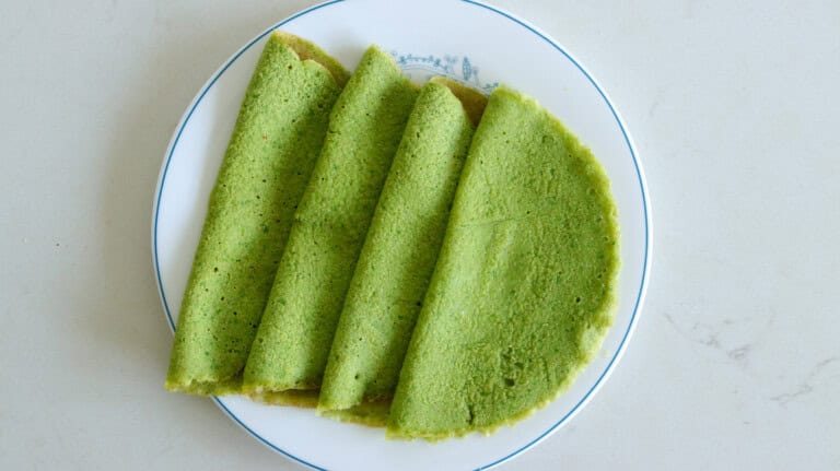A white plate with four folded green spinach crepes arranged in a fan shape on a light-colored surface.
