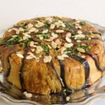 A round maqluba dish is plated on a silver serving platter. The dish is wrapped in thin slices of roasted eggplant and topped with a garnish of chopped herbs and sliced almonds.