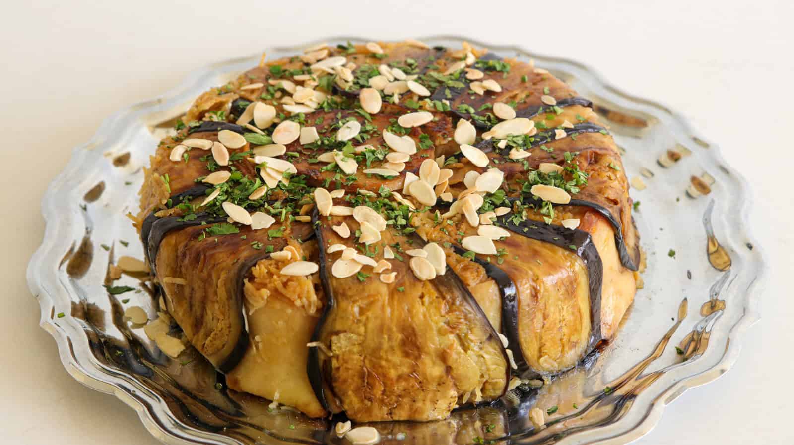 A round, savory dish is plated on a silver serving platter. The dish is wrapped in thin slices of roasted eggplant and topped with a garnish of chopped herbs and sliced almonds.