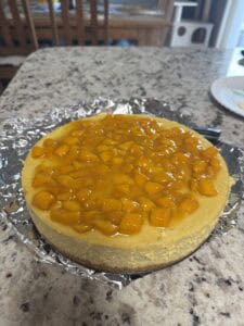 A cheesecake topped with glossy diced mango pieces sits on a foil-covered base on a granite countertop, with wooden chairs and a plate visible in the background.