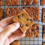 A hand is holding a square peanut butter banana bar with chocolate chips. More bars are arranged on a cooling rack in the background. The text "Peanut Butter Banana Bars" is displayed at the top.