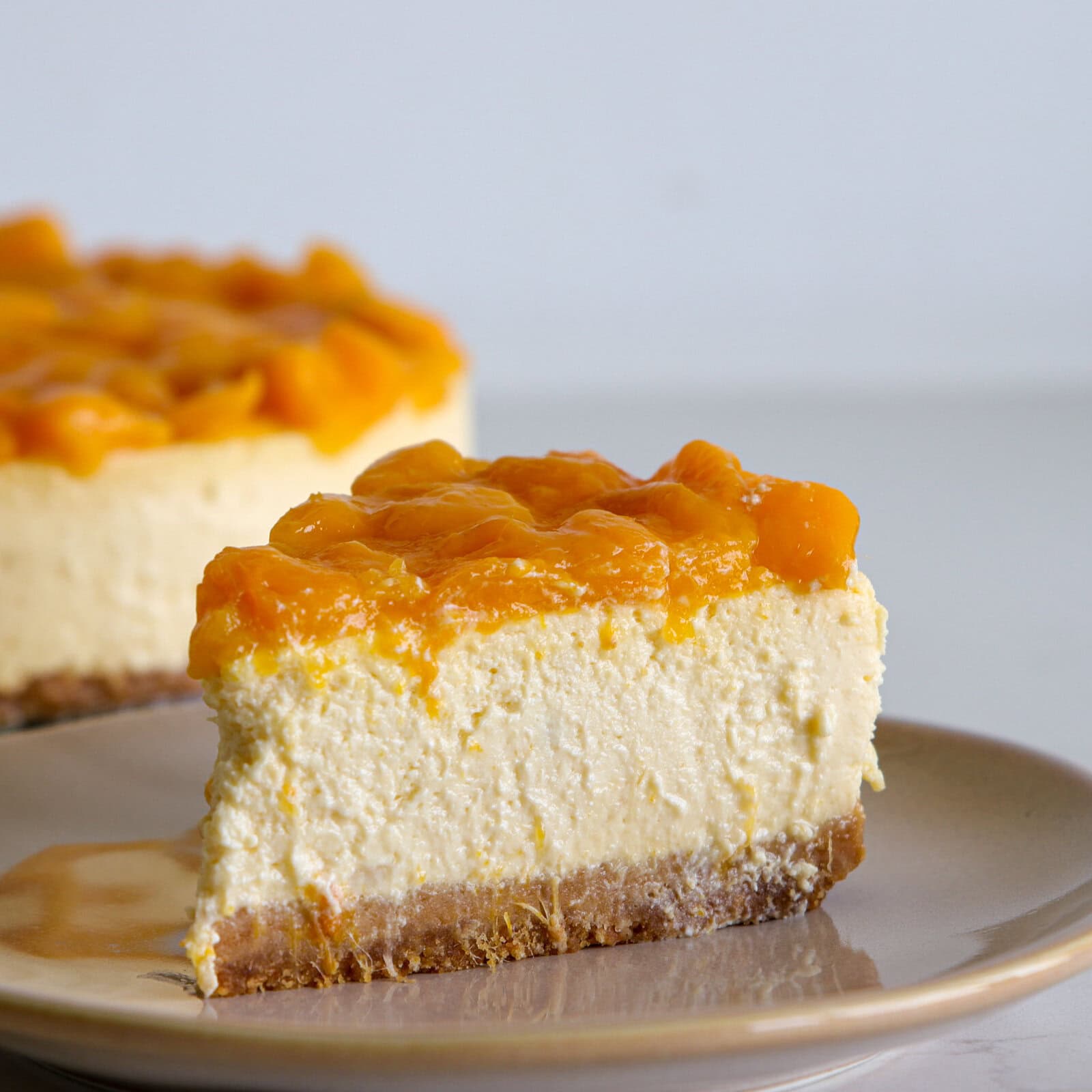 A slice of cheesecake topped with a layer of mango chunks sits on a beige plate, with the rest of the cheesecake visible in the background.