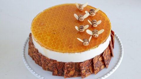 A round cake with a honeycomb-patterned topping and adorned with small, bee-shaped decorations made of fondant or marzipan. The sides of the cake are lined with honeycomb-like edible pieces, and the cake sits on a white plate. The background is plain, light-colored.