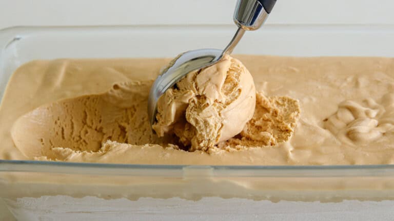 A metal ice cream scooper is scooping a creamy ball of coffee-flavored ice cream from a rectangular glass container filled with the same ice cream. The background is a light, neutral color.