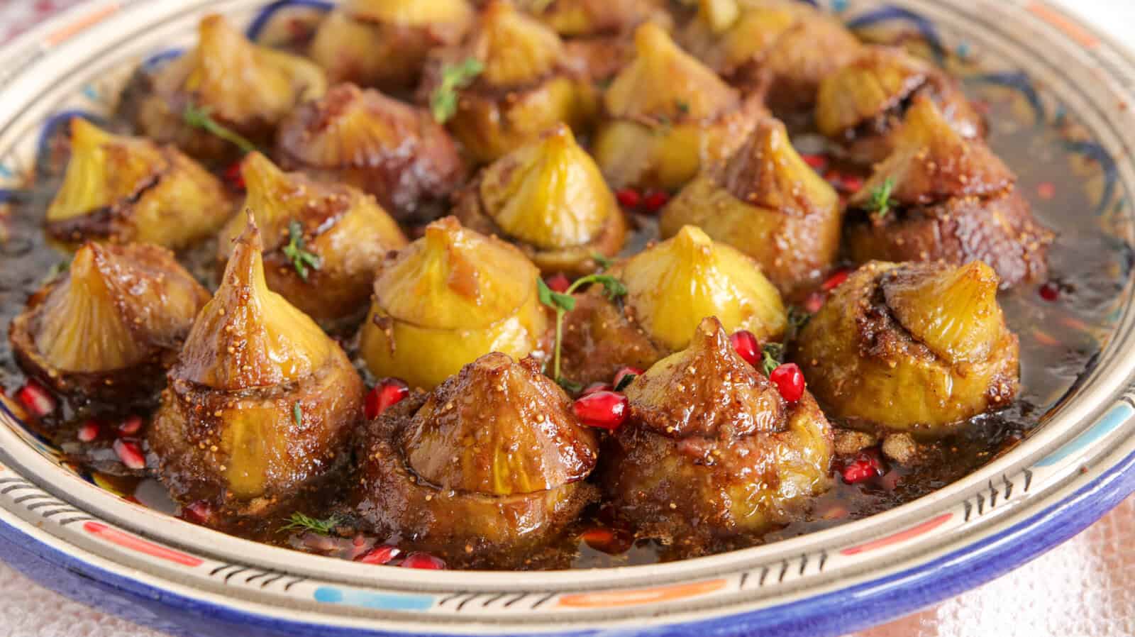 A colorful plate filled with meat-stuffed figs in a rich, brown sauce, garnished with pomegranate seeds and herbs. The onions are arranged upright and closely packed, showcasing their golden tops.