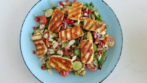 A fresh salad topped with grilled halloumi cheese, cherry tomatoes, cucumber slices, pomegranate seeds, almond flakes, and a mix of greens on a light blue plate. The grilled cheese has distinct grill marks and rests atop the vibrant vegetables.