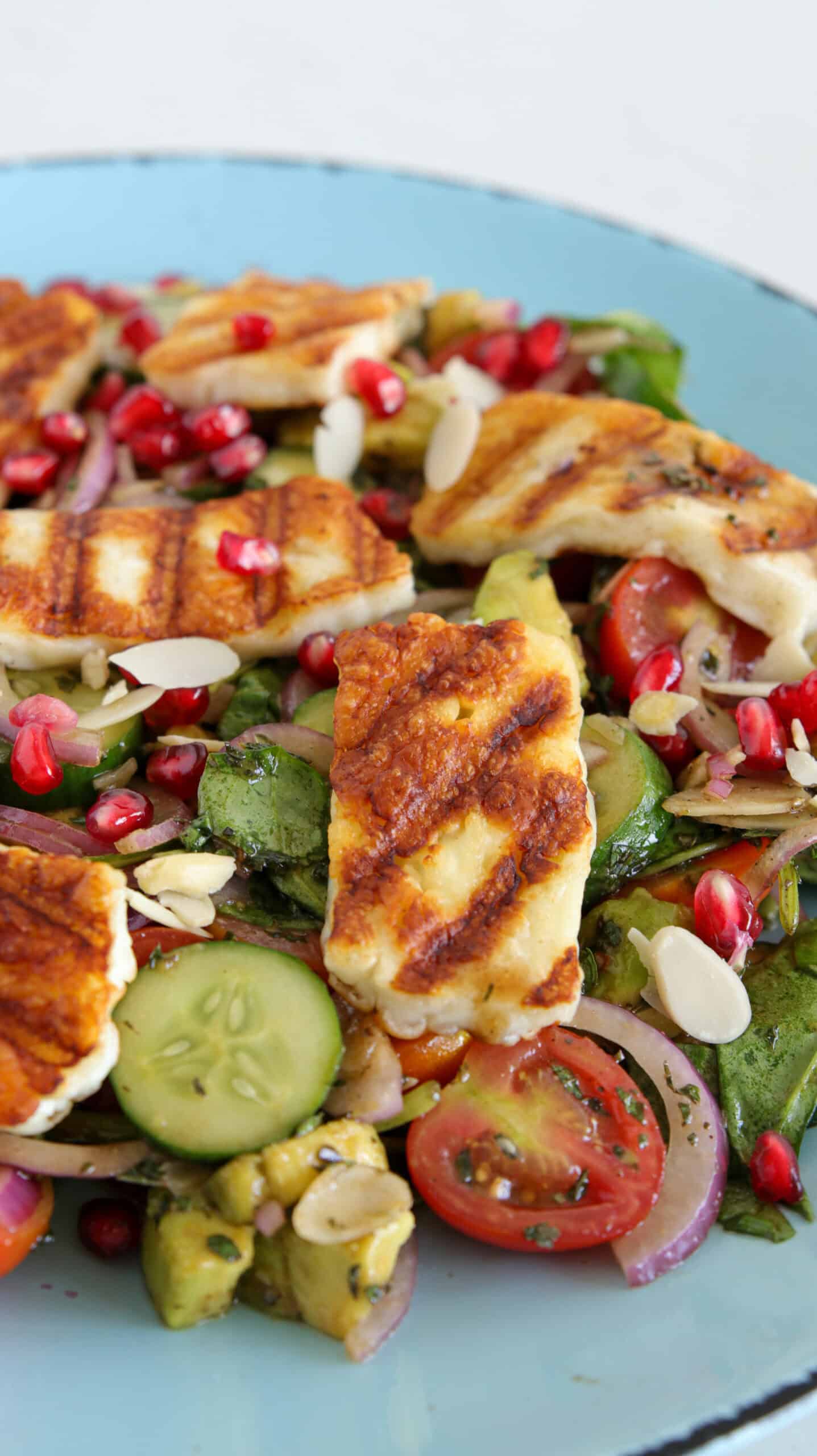 A fresh salad topped with grilled halloumi cheese, cherry tomatoes, cucumber slices, pomegranate seeds, almond flakes, and a mix of greens on a light blue plate. The grilled cheese has distinct grill marks and rests atop the vibrant vegetables.