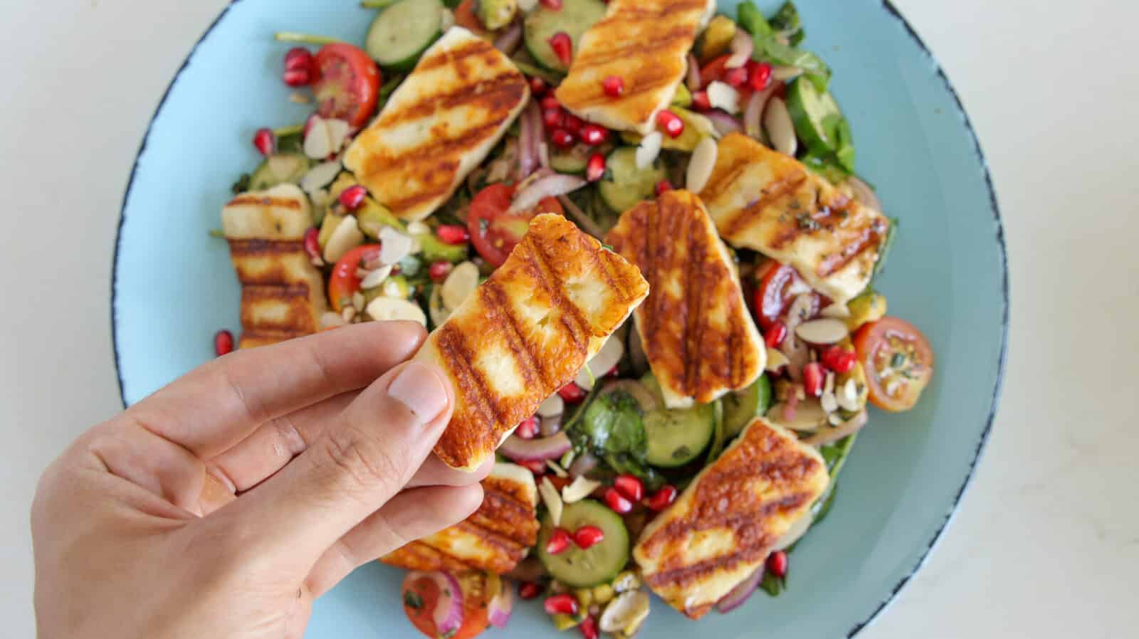 a hand holding a slice of grilled halloumi cheese over a salad