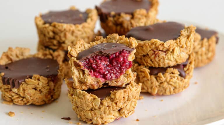 A close-up of stacked oat cups filled with a dark chocolate topping and a raspberry jam center. One of the cups has a bite taken out, revealing its vibrant raspberry filling. The oat cups are placed on a white surface.