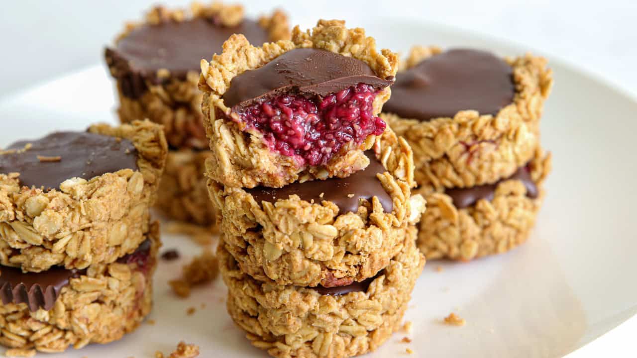 A close-up of stacked oat cups filled with a dark chocolate topping and a raspberry jam center. One of the cups has a bite taken out, revealing its vibrant raspberry filling. The oat cups are placed on a white surface.