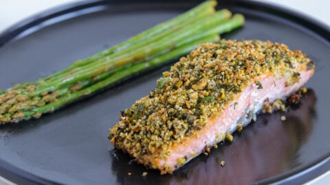 A serving of baked salmon fillet topped with a golden, crispy herb crust is plated alongside a bundle of tender asparagus spears. Both are presented on a dark, round plate.