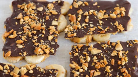 A rectangular dessert topped with smooth melted chocolate and sprinkled with chopped nuts on a white surface. The edges of the dessert show glimpses of a light, flaky pastry underneath the chocolate layer.