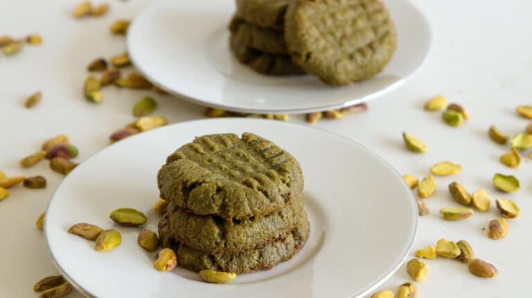 A stack of pistachio cookies sits on a white plate with scattered pistachios nearby. Another plate with a stack of cookies is in the background. The text "PISTACHIO COOKIES" and "4-INGREDIENTS" is overlaid on the image.