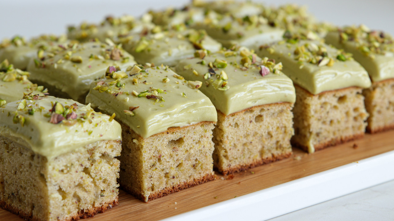 Sliced pieces of moist cake with a pale green frosting, topped with chopped pistachios. The cake rests on a wooden surface and appears to have a light, fluffy texture.