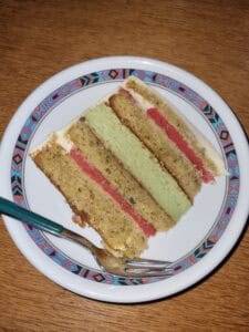 A slice of layered cake with green and pink filling on a decorative white plate, placed on a wooden table. A fork is resting on the plate beside the cake.