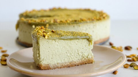 A rich green pistachio cheesecake topped with crushed nuts on a white cake stand. A slice has been lifted from the cake, showcasing its creamy texture and a light brown crust.