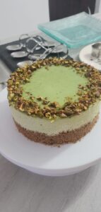 A round, layered pistachio cake with a green top, decorated with chopped pistachios around the edge, sits on a white plate on a kitchen counter. Baking utensils and glassware are in the background.