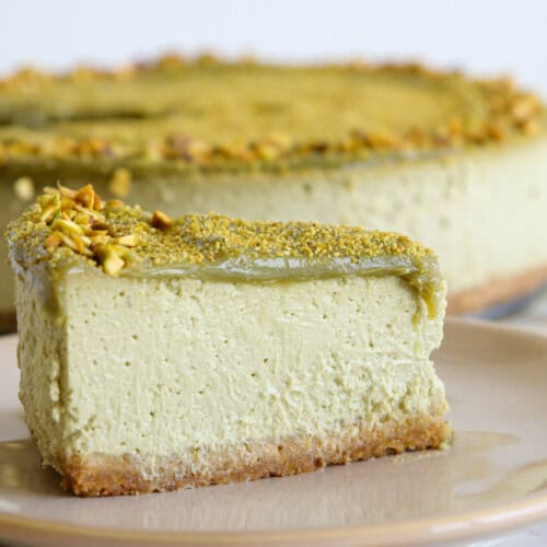 A slice of pistachio cheesecake on a beige plate with crushed pistachios sprinkled on top, and the rest of the cheesecake in the background. Some pistachio pieces are scattered on the white surface.