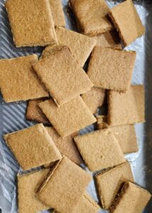 A pile of square, golden-brown biscuits or cookies is spread on a silver tray with a ridged pattern. The biscuits appear baked and have a slightly rough texture.