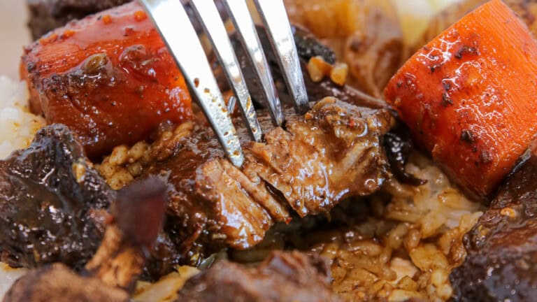 A close-up of a fork pulling apart tender braised beef with chunks of carrot on a bed of rice, all coated in a rich brown sauce.