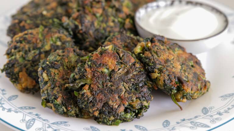 A plate featuring several golden-brown spinach fritters, served alongside a small bowl of creamy white dip. The fritters have a slightly crispy exterior and are placed on a white plate with blue floral patterns.