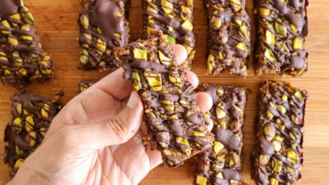 A close-up of several granola bars topped with chopped pistachios and drizzled with chocolate, arranged neatly on a wooden surface. The bars have a rich, textured appearance and the green pistachios contrast with the dark chocolate.