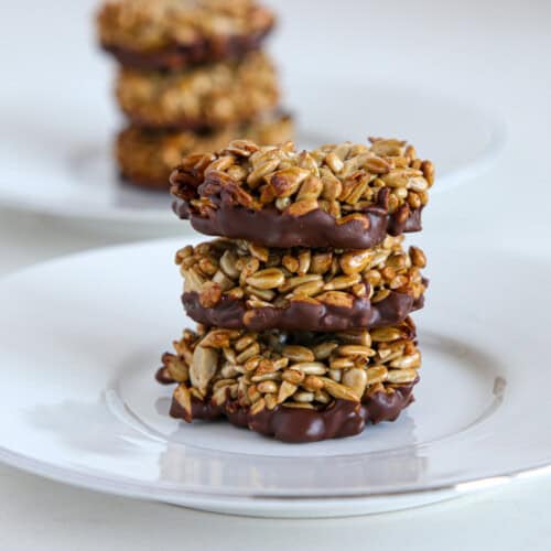 Three homemade sunflower seed and chocolate clusters are stacked on a white plate in the foreground, while additional clusters are visible on a similar plate in the background. The clusters appear to be bound together with a sweet, sticky coating.