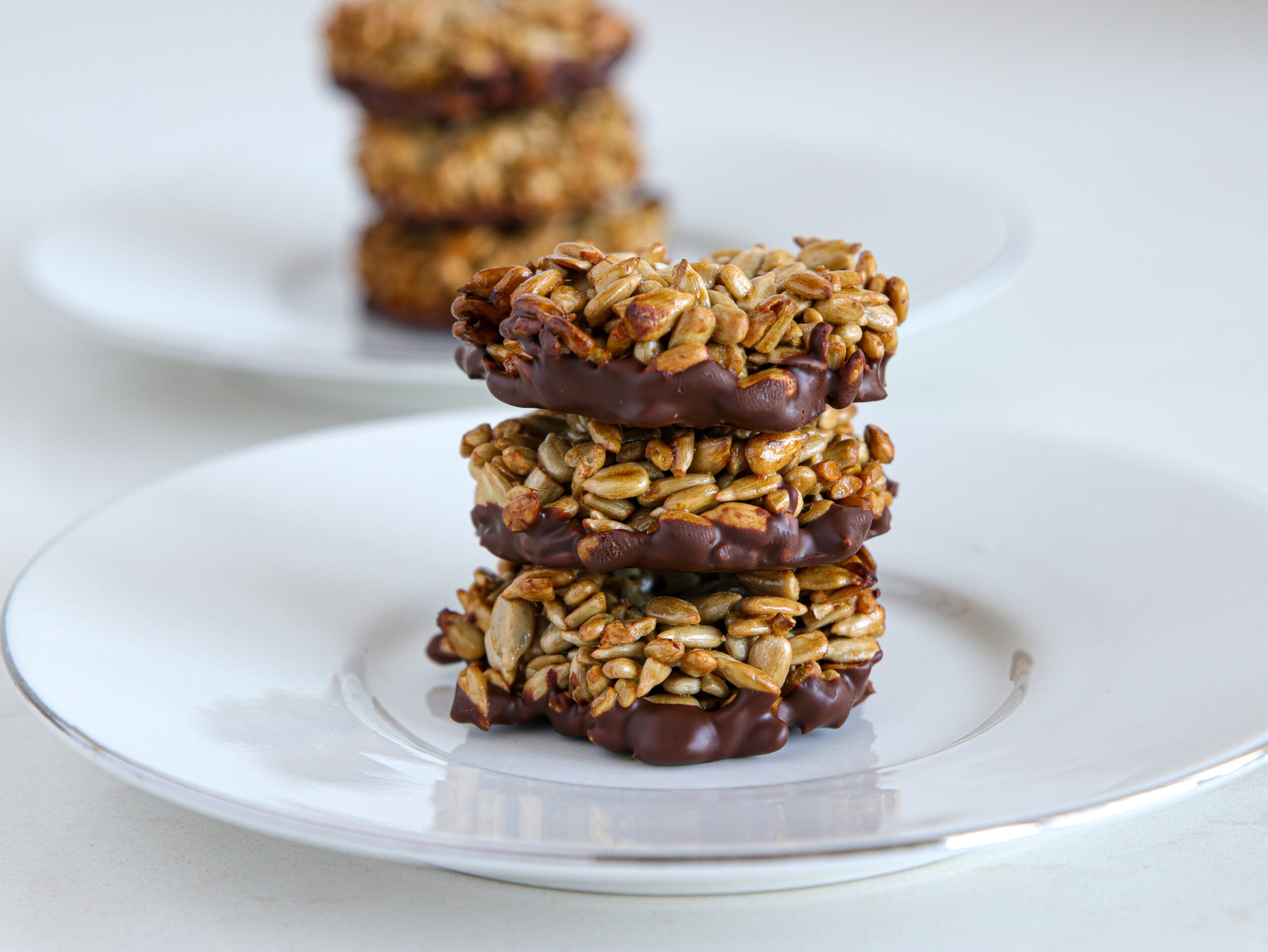 Three homemade sunflower seed and chocolate clusters are stacked on a white plate in the foreground, while additional clusters are visible on a similar plate in the background. The clusters appear to be bound together with a sweet, sticky coating.