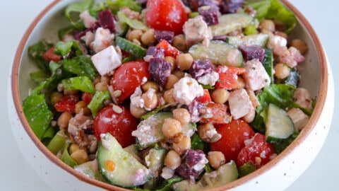 A colorful Greek salad in a bowl, featuring cherry tomatoes, cucumber slices, chopped bell peppers, chickpeas, olives, feta cheese chunks, and mixed greens, all tossed in a creamy dressing.