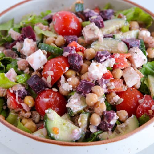 A bowl of fresh salad featuring mixed greens, cherry tomatoes, diced cucumbers, chickpeas, feta cheese, and pieces of beetroot. The colorful ingredients are tossed together in a white, speckled ceramic bowl.