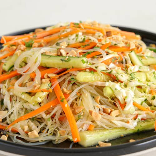 A black plate is filled with a colorful Asian noodle salad. The salad includes rice noodles, julienned carrots, cucumber slices, bean sprouts, and chopped peanuts, all topped with a light dressing and garnished with fresh herbs.