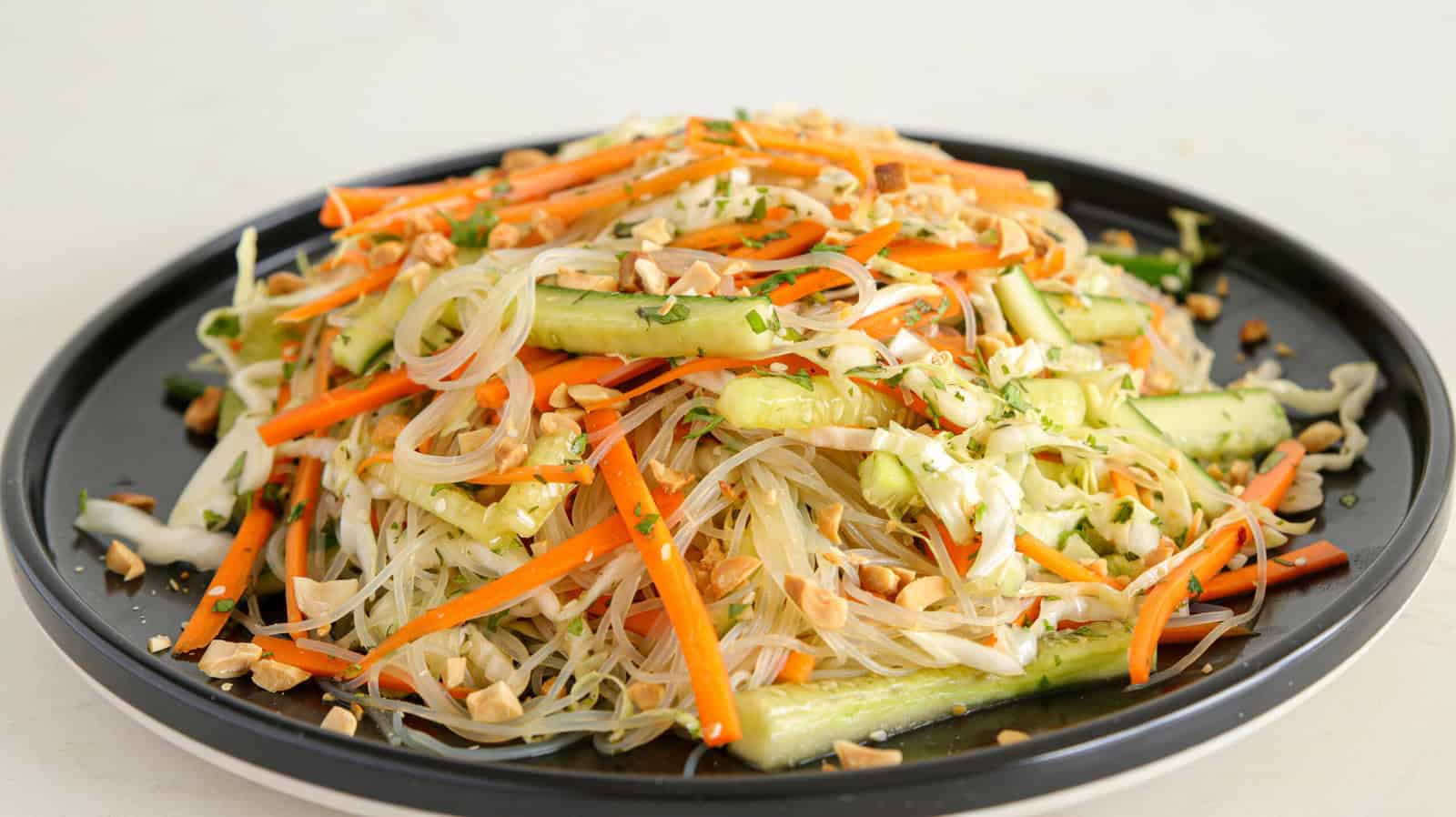 A black plate is filled with a colorful Asian noodle salad. The salad includes rice noodles, julienned carrots, cucumber slices, bean sprouts, and chopped peanuts, all topped with a light dressing and garnished with fresh herbs.