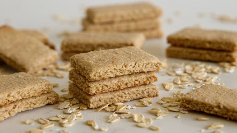 Stacked and scattered rectangular oat biscuits lie on a light surface, surrounded by a sprinkling of uncooked oats.