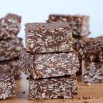 Several pieces of chocolate coconut squares are neatly stacked on a wooden surface. The squares have a rich, dark chocolate color with visible shredded coconut pieces mixed in. The background is simple and out of focus, emphasizing the texture of the treats.